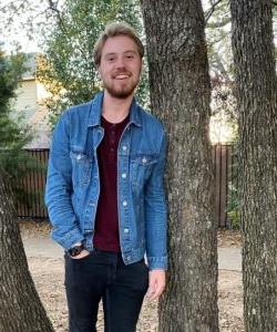 A man in a denim jacket and dark pants smiling between two trees on Howard Payne University campus. | HPU